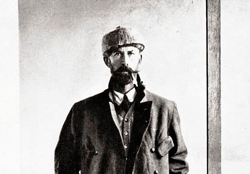 Portrait en noir et blanc de Percy Fawcett, célèbre explorateur britannique du début du XXe siècle. Il est debout sur une véranda en bois, vêtu d'un long manteau de style militaire, d’un pantalon de toile, de bottes montantes et d’une casquette en laine