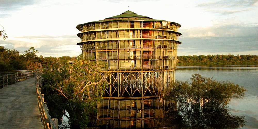 Grande tour circulaire en bois, construite sur pilotis au-dessus de l’eau, illuminée par les teintes dorées du coucher du soleil dans la forêt amazonienne.