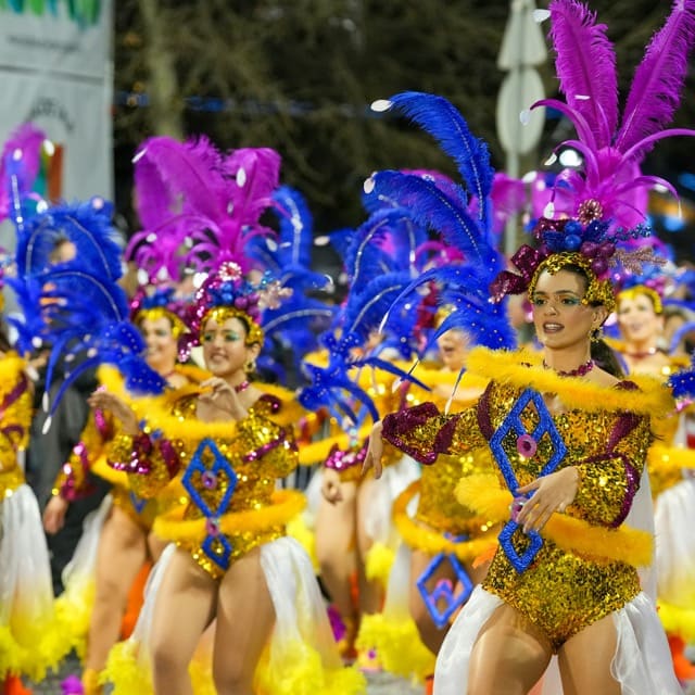 Groupe de danseuses en pleine parade carnavalesque, vêtues de costumes scintillants dorés agrémentés de plumes bleues et roses, de strass et de motifs colorés.