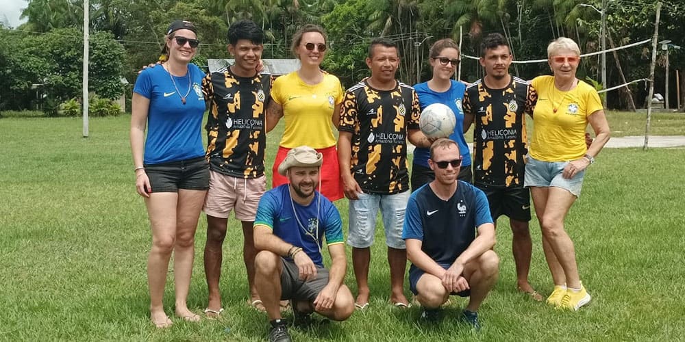 Photo de groupe prise sur une pelouse au cœur d’un village amazonien : cinq joueurs locaux portant un maillot noir et jaune Heliconia, entourés de voyageurs vêtus de t-shirts bleus ou jaunes. 