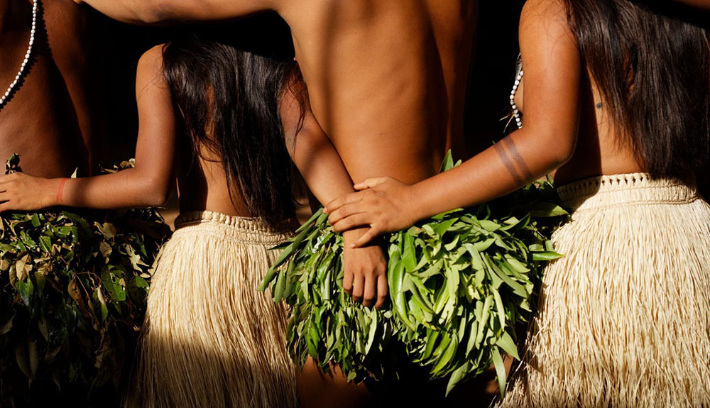 Plan rapproché des hanches et dos de quatre danseurs autochtones enlacés : jupes de fibres naturelles et jupons de feuilles vertes, longues chevelures noires, bracelets de perles et bras entrecroisés, illustrant l’unité du groupe lors d’une célébration traditionnelle.