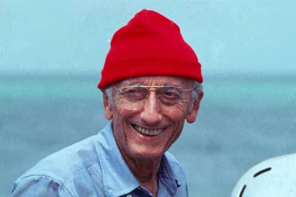 Jacques-Yves Cousteau souriant, portant un bonnet rouge et des lunettes, vêtu d'une chemise bleue.