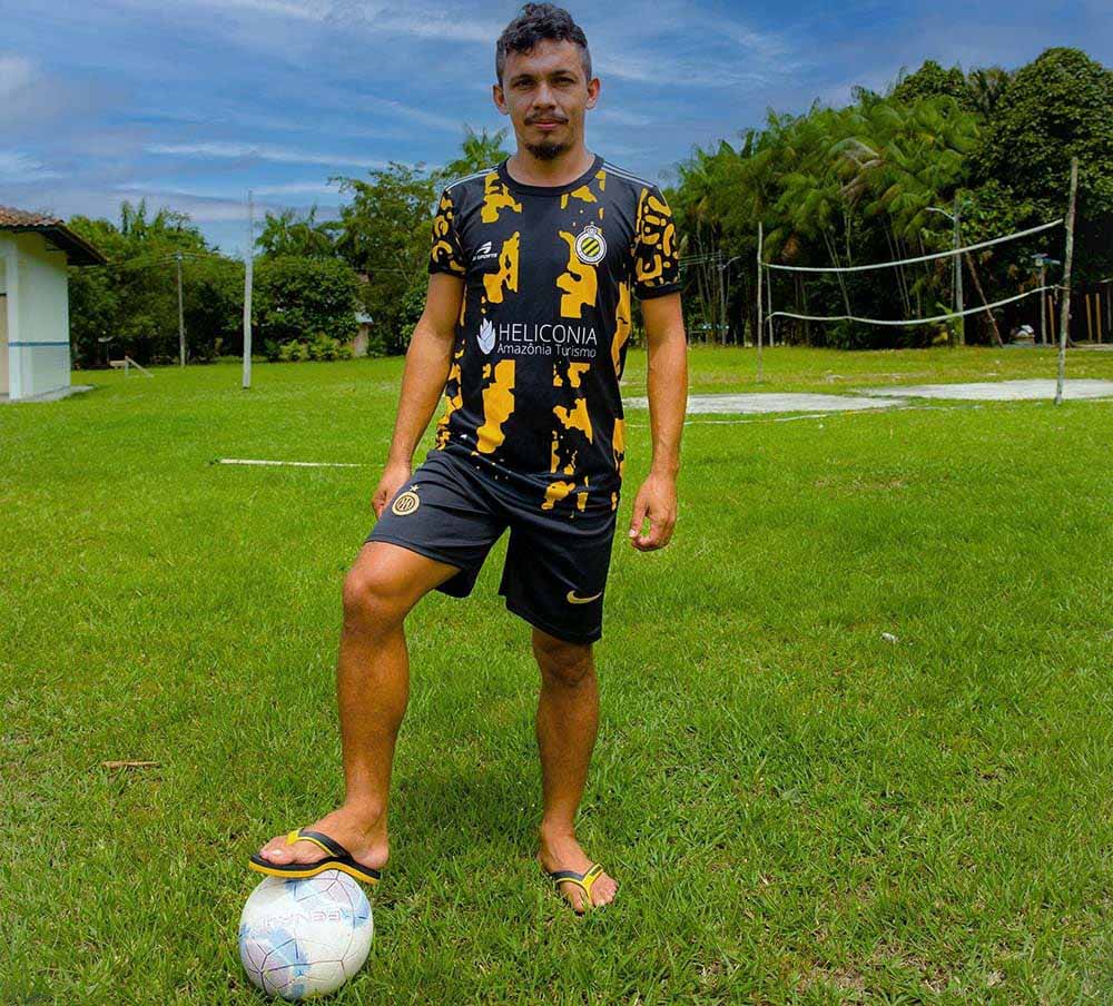 Jeune joueur debout sur une pelouse verte, un pied posé sur un ballon. Il porte le maillot noir et jaune Heliconia et un short noir Nike, ainsi que des tongs. 