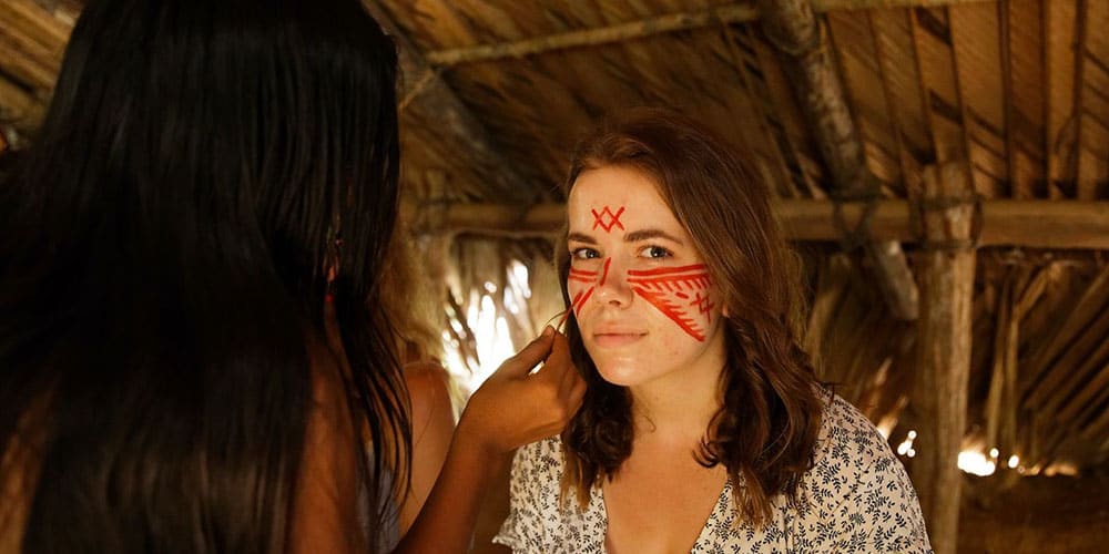Une voyageuse assise se fait tracer des motifs rouges au roucou sur le visage par une femme de la communauté tatuyo, dans une maloca traditionnelle
