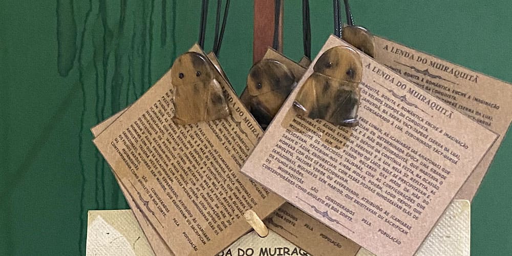Plusieurs amulettes en forme de grenouille, appelées Muiraquitã, sont accrochées à un présentoir avec une fiche expliquant leur légende.