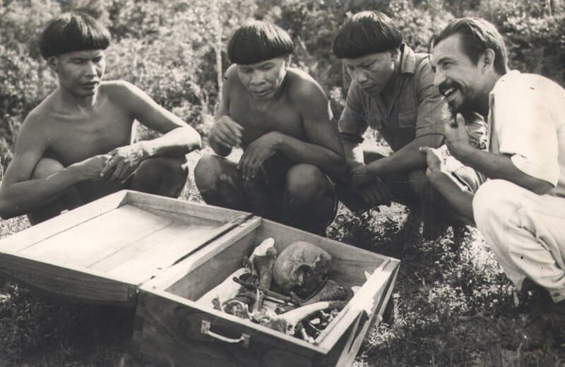 Photographie en noir et blanc montrant Orlando Villas-Bôas, un célèbre explorateur et défenseur des peuples autochtones du Brésil, accroupi aux côtés de trois hommes autochtones Xingu. Tous observent une caisse ouverte contenant des ossements humains