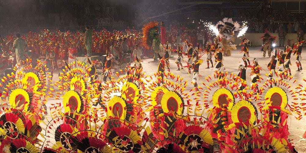 Une vaste parade de danseurs et danseuses en costumes indigènes colorés défile dans une arène éclairée, avec de grandes coiffes à plumes jaunes, rouges et noires.