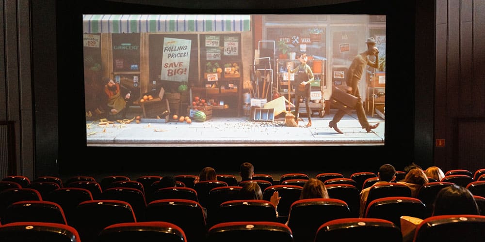 ntérieur d’une salle de cinéma aux fauteuils rouges presque vides ; quelques spectateurs sont assis devant un grand écran où passe un film d’animation montrant une scène de rue.