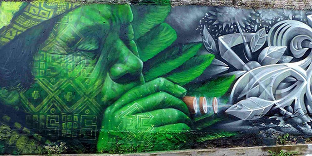 Portrait mural d’un visage chamanique en vert, yeux fermés, soufflant à travers un tuyau ou une pipe.