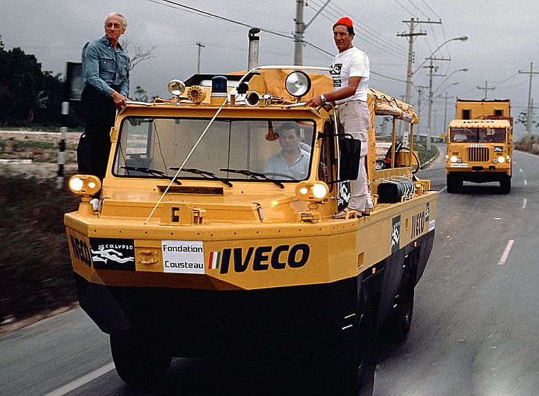 Véhicule amphibie jaune de la Fondation Cousteau sur une route, avec deux hommes à bord, dont l’un porte un bonnet rouge.