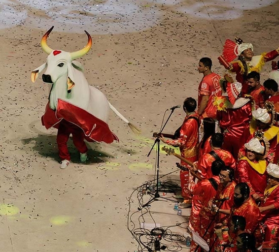 Un personnage déguisé en taureau blanc décoré de rouge danse devant un groupe de musiciens en costumes rouges ornés de dorures