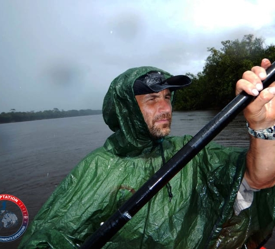 Un homme barbu, vêtu d’un poncho imperméable vert, pagaie sous une forte pluie sur un fleuve amazonien. 