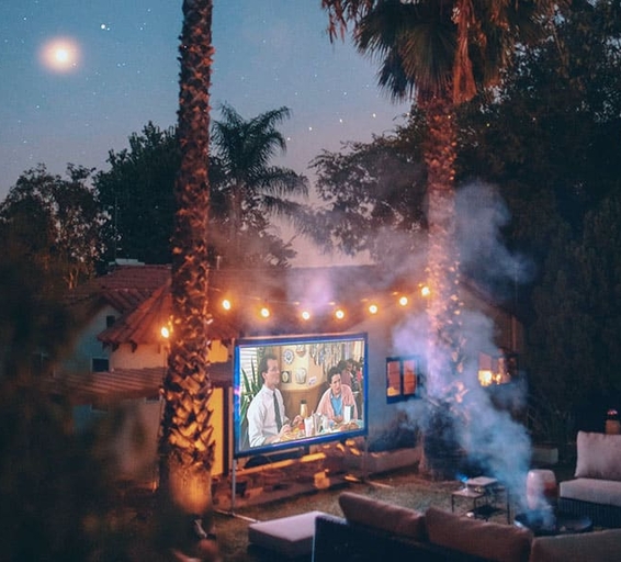 Jardin aménagé pour une projection nocturne : un écran lumineux diffuse un film tandis que des guirlandes d’ampoules suspendues entre les palmiers créent une atmosphère chaleureuse. 