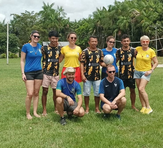 Photo de groupe prise sur une pelouse au cœur d’un village amazonien : cinq joueurs locaux portant un maillot noir et jaune Heliconia, entourés de voyageurs vêtus de t-shirts bleus ou jaunes. 