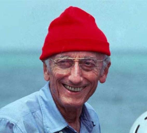 Jacques-Yves Cousteau souriant, portant un bonnet rouge et des lunettes, vêtu d'une chemise bleue.